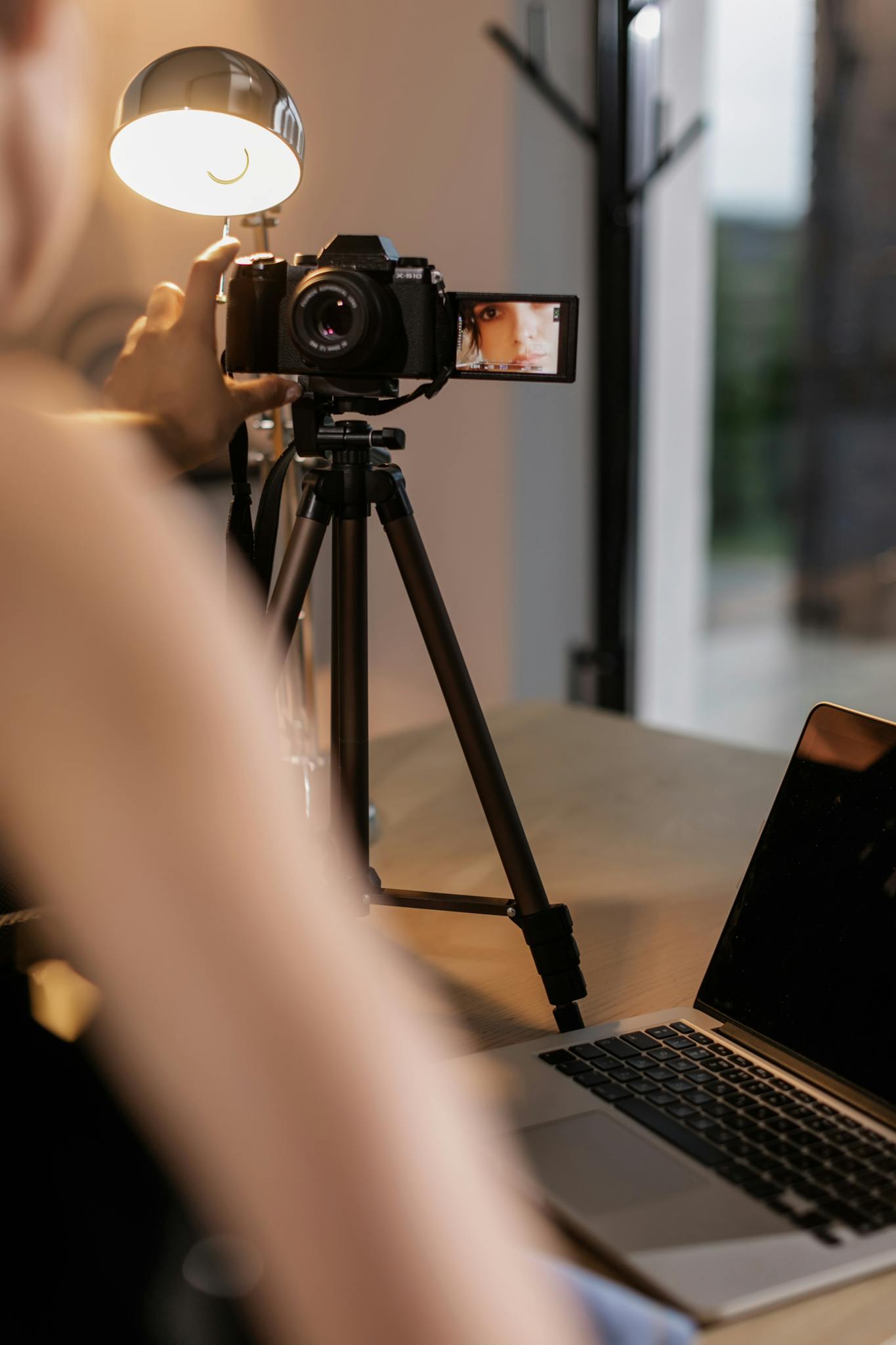 A person sets up a DSLR camera on a tripod for vlogging indoors, illuminated by a lamp.